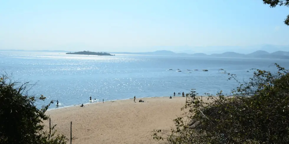Imagem da Ilha de Paquetá em um dia de céu aberto. Mostra a areia , o mar e algumas pessoas na beira.
