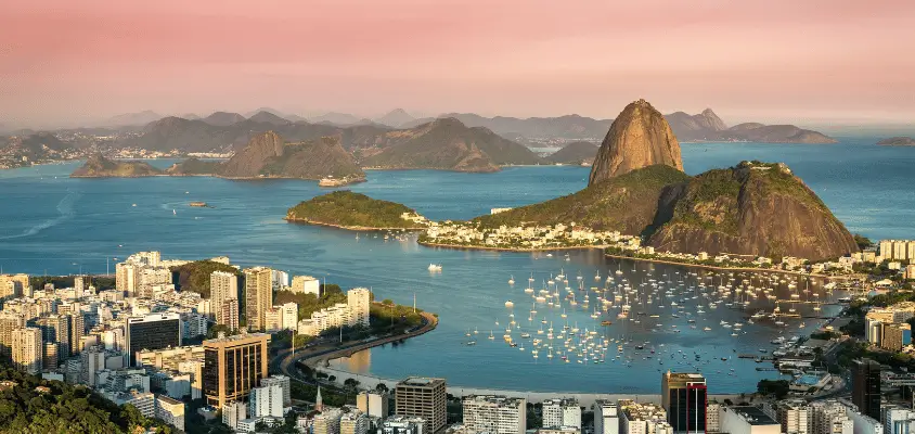 Imagem panorâmica da cidade do Rio de Janeiro no entardecer, mostrando a Baía de Guanabara, parte da cidade e o Pão de Açúcar.