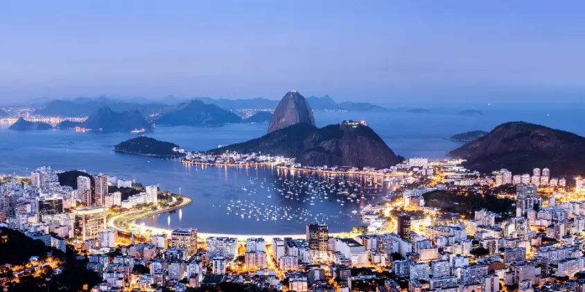 Foto panorâmica do Rio de Janeiro no início da noite. Ao fundo está o Pão de Açúcar e a Baía de Guanabara