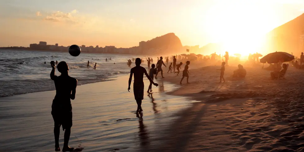 Muitas pessoas na praia de Ipanema com o sol caindo. No primeiro plano, dois meninos jogando futebol na areia